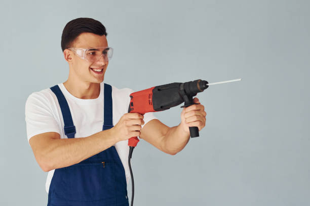 istock In protective eyewear and with drill in hands. Male worker in blue uniform standing inside of studio against white background 1330863671