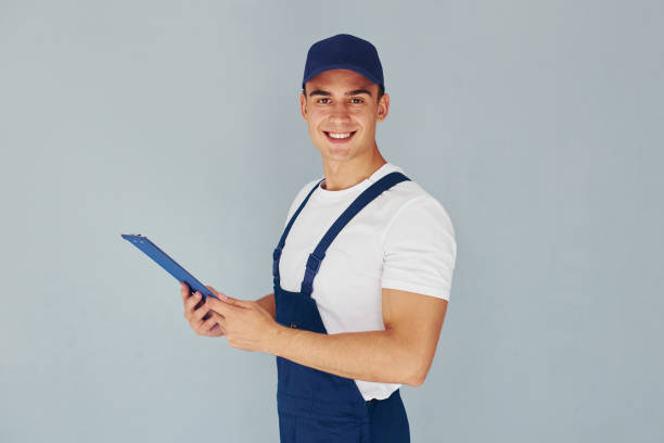 istock In cap and with notepad. Male worker in blue uniform standing inside of studio against white background 1330863835