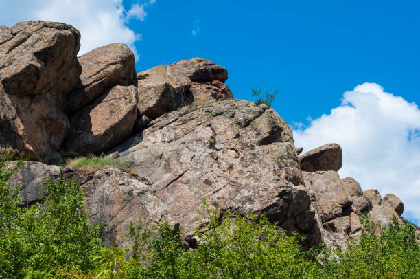 istock Granite rocks close-up on background blue sky with white clouds, green leaves on treetops. 1330864189