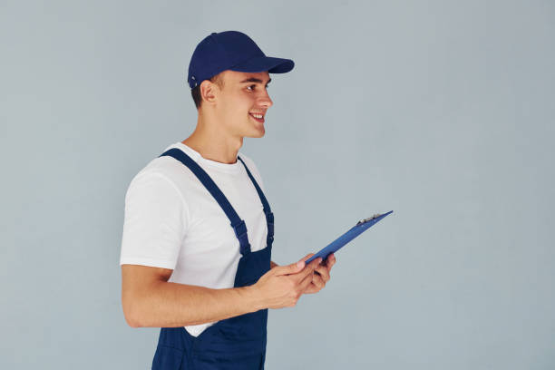 istock In cap and with notepad. Male worker in blue uniform standing inside of studio against white background 1330864814