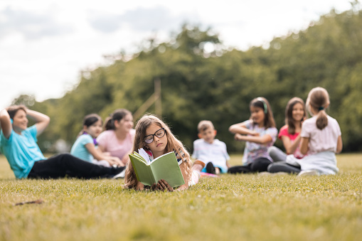 Schoolgirl reading a book while her friends having fun playing in the nature
