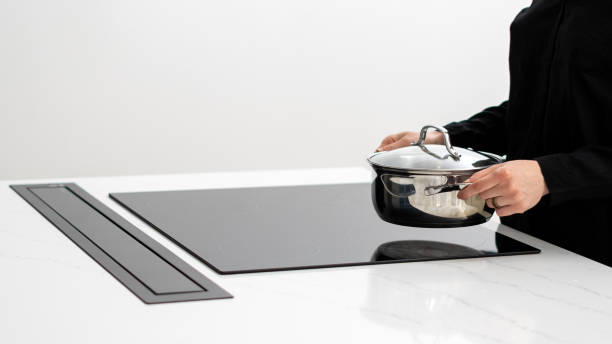 Woman putting metal pot on induction stove Woman in black shirt holding metal pot, going to put it on built in glass induction stove while cooking food in white kitchen with integrated technology, cropped shot glass ceramic stove top stock pictures, royalty-free photos & images