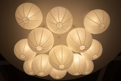 Larger number of large, white paper ball lampshades with lighting switched on. against a dark background