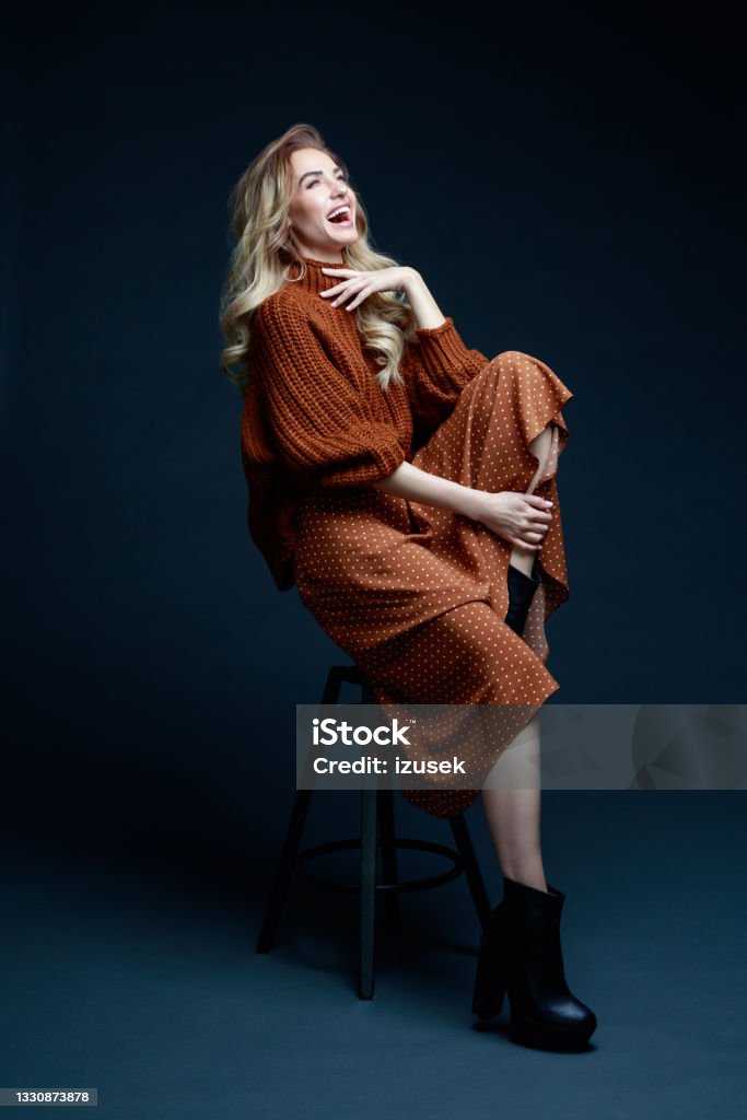 Fashion portrait of elegant woman in brown clothes, dark background Portrait of long hair blond young woman wearing brown sweater and skirt, sitting on chair, looking away and laughing. Studio shot against black background. Fashion Model Stock Photo
