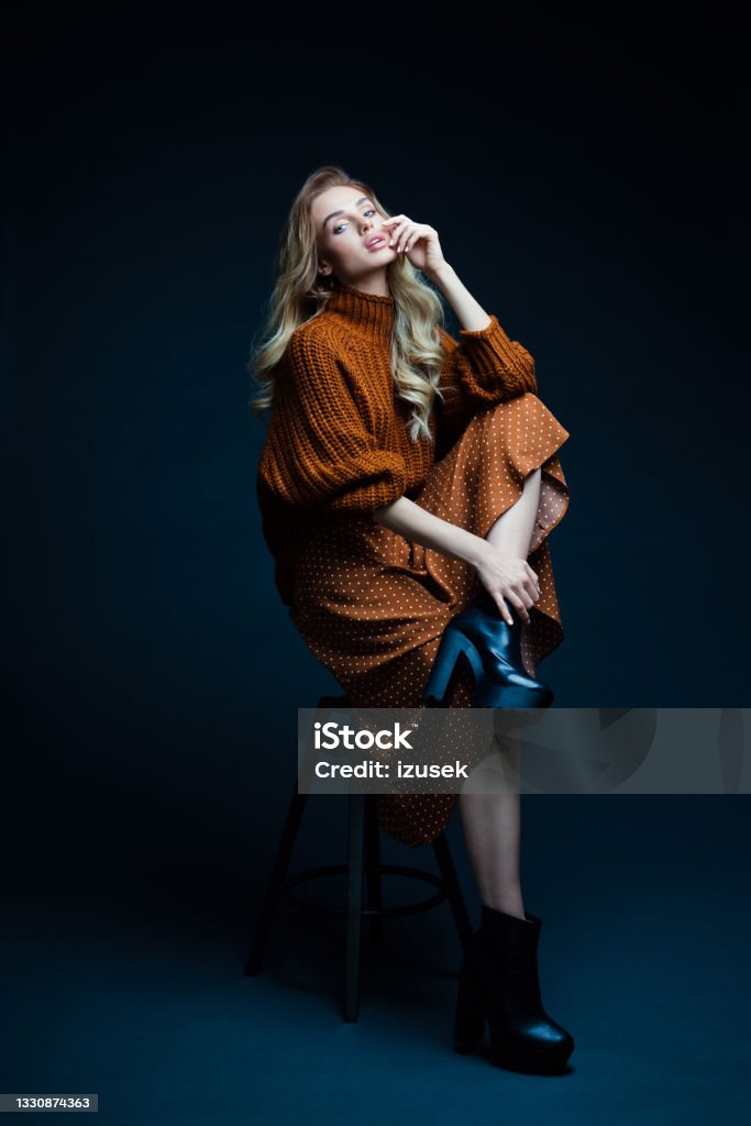Fashion portrait of elegant woman in brown clothes, dark background Portrait of long hair blond young woman wearing brown sweater and skirt, sitting on chair, looking at camera. Studio shot against black background. Fashion Model Stock Photo