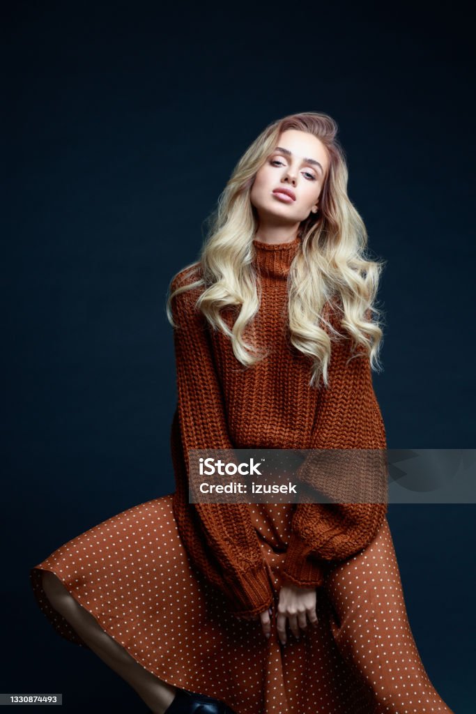 Fashion portrait of elegant woman in brown clothes, dark background Portrait of long hair blond young woman wearing brown sweater and skirt, sitting on chair, looking at camera. Studio shot against black background. Women Stock Photo