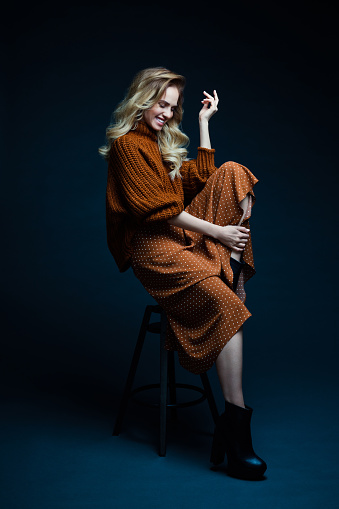 Portrait of long hair blond young woman wearing brown sweater and skirt, sitting on chair, looking away and laughing. Studio shot against black background.