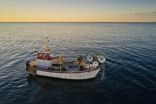 Full length view from drone perspective of mid adult Caucasian man standing on deck of small trawler preparing his nets with horizon over water in background.