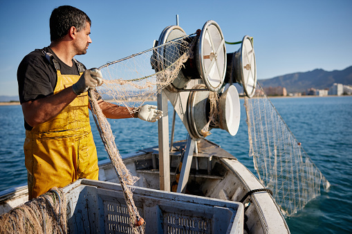 Mid adult Caucasian man in t-shirt and bib overalls standing at bow of small trawler and managing net while at sea in Mediterranean.