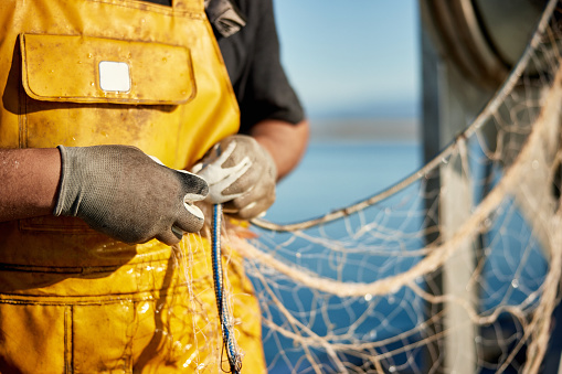 Close-up of Caucasian man in bib overalls and gloves standing in sunshine on deck of fishing boat organizing net with calm Mediterranean in background.