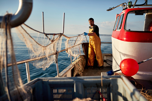 Full length view of Caucasian man in protective workwear standing at stern of boat managing nets while at sea in early morning.