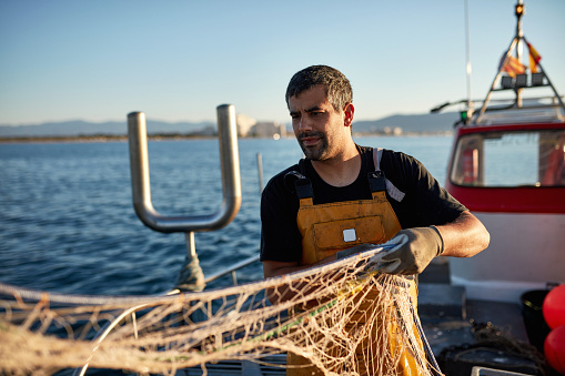 Waist-up front view of Caucasian man in t-shirt and bib overalls working on deck of small trawler in Mediterranean Sea.