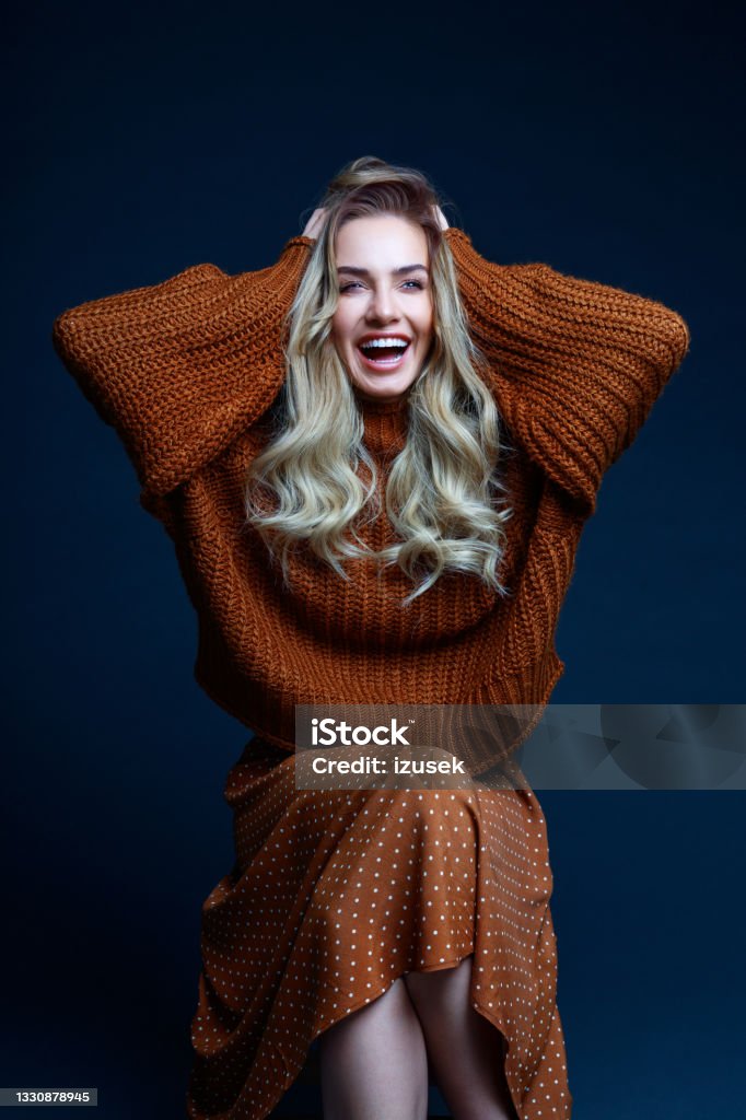 Fashion portrait of cheerful woman in brown clothes, dark background Portrait of long hair blond young woman wearing brown sweater and skirt, sitting on chair, laughing at camera. Studio shot against black background. Fashion Stock Photo
