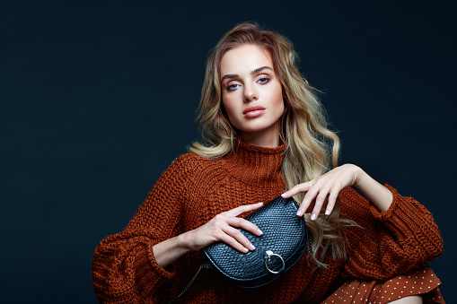 Portrait of long hair blond young woman wearing brown sweater and skirt, holding black purse and looking at camera. Studio shot against black background.