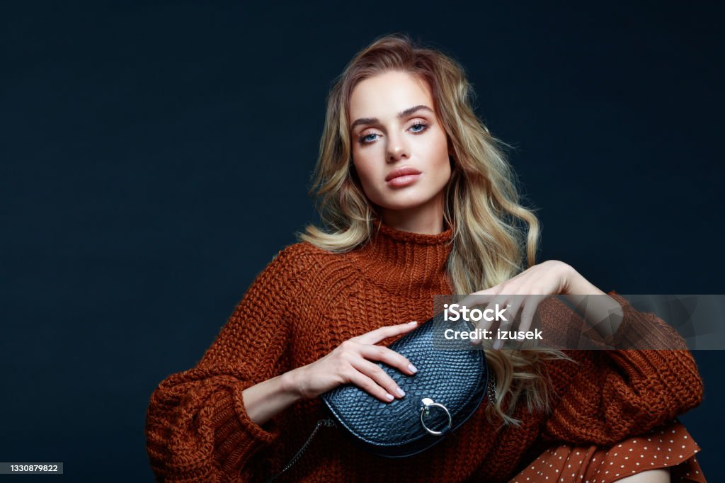 Fashion portrait of elegant woman in brown clothes, dark background Portrait of long hair blond young woman wearing brown sweater and skirt, holding black purse and looking at camera. Studio shot against black background. Luxury Stock Photo