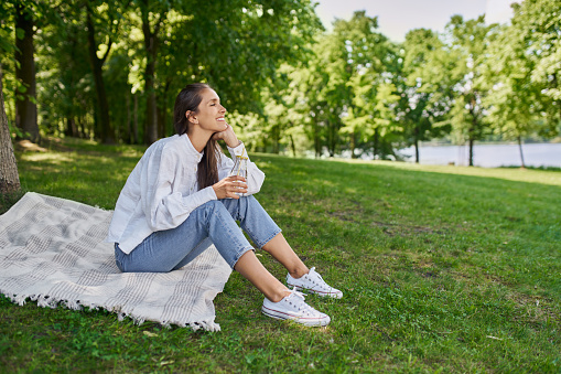 Smiling young woman sitting on blanket in park drinking water during beautiful summer day