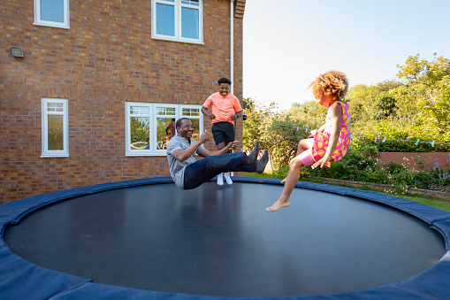 A young girl and her brother jumping on a trampoline outdoors in the back garden at home with their grandfather. They are all trying to do a seat drop jump.