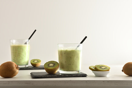 Two glasses of kiwi drink and containers with cut fruit around on table isolated white. Front view.