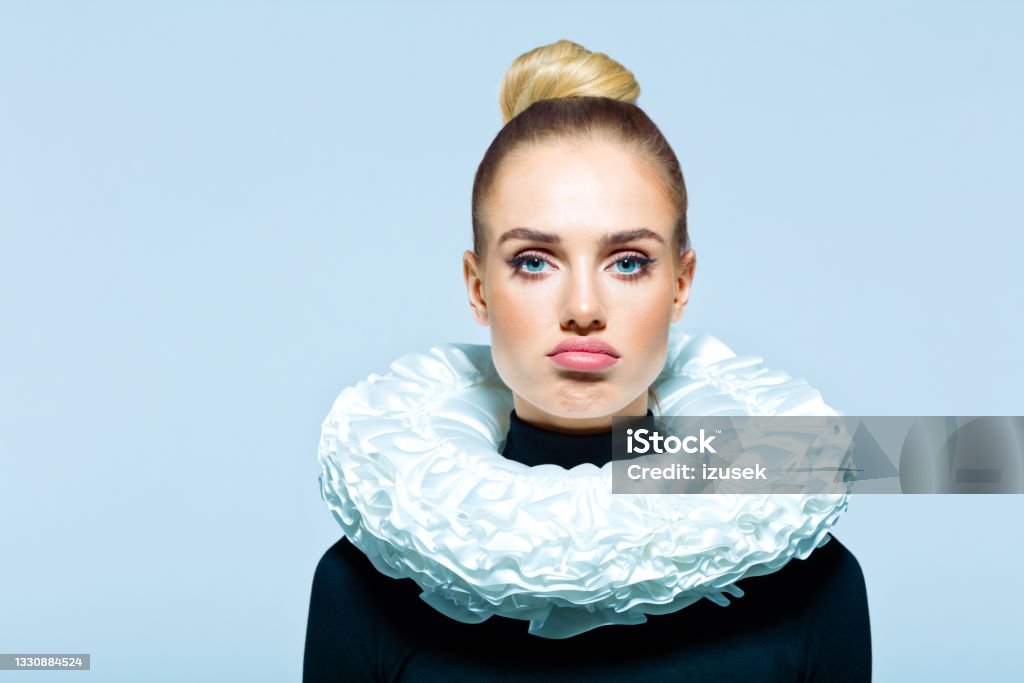Portrait of woman wearing neck ruff Disappointed woman wearing black turtleneck and white neck ruff looking at camera. Studio shot on blue background. Women Stock Photo