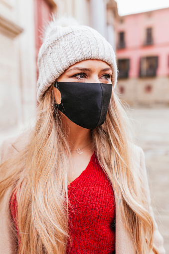 Portrait of young blond latin woman in stylish coat and woolen hat wearing protective mask in an old street