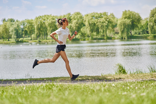 Side view of young fit woman running in park during sunny summer day