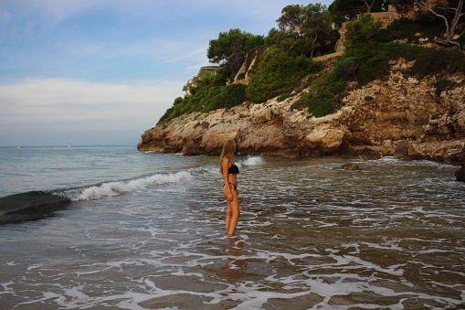 Salou. Spain - 09 September 2018. A blonde girl with loose hair stands in a half-turn in a swimsuit on the beach. on the background of a rock with trees.