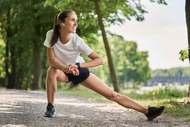 runner donna sorridente che si estende nel parco - pantaloncini da corsa foto e immagini stock