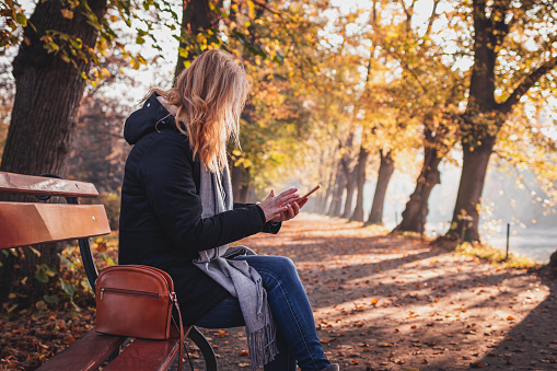 Woman sitting on bench in autumn park and using smart phone. Relaxation and text messaging on mobile phone during city break in fall season