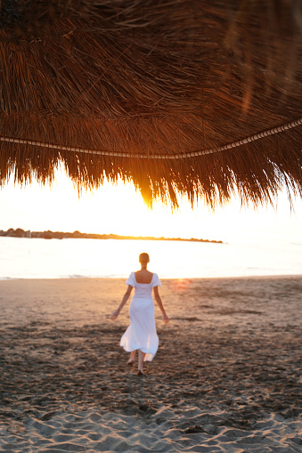 Back view of woman running in white maxi dress on the beach at sunrise. Freedom. Real people. Vintage style.