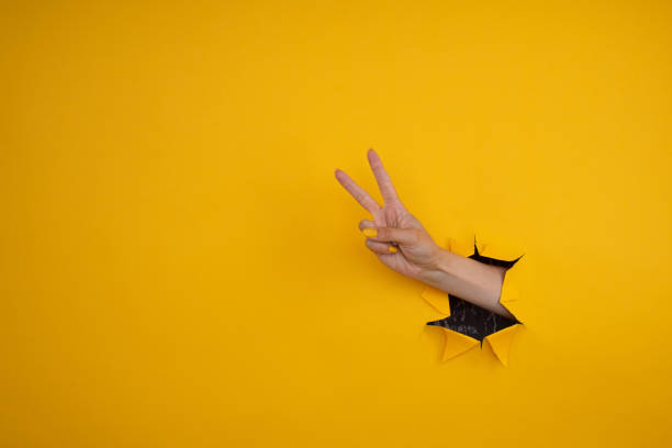 istock Hand showing peace gesture through torn yellow paper background. Victory, V sign. 1330895611