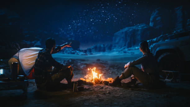 istock Lovely Young Couple Sitting by Campfire Watching Night Sky while Camping in the Canyon. Girlfriend and Boyfriend Tell Inspirational Nature Loving Stories about Life, Look at Milky Way Stars 1330897018