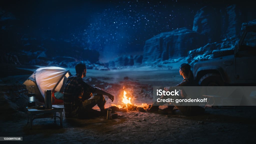 Happy Couple Sitting by Campfire Watching Night Sky while Camping in the Canyon. Two Traveling people Tell Inspirational Stories at Campsite, Look at Milky Way Stars. On Holiday Vacation Trip Camping Stock Photo