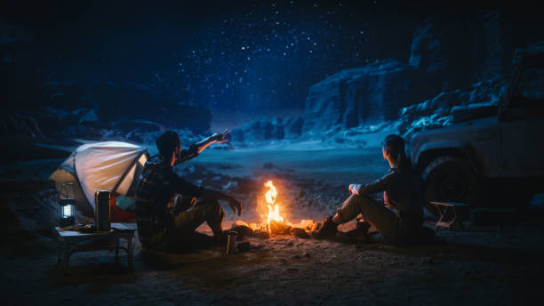 istock Lovely Young Couple Sitting by Campfire Watching Night Sky while Camping in the Canyon. Girlfriend and Boyfriend Tell Inspirational Nature Loving Stories about Life, Look at Milky Way Stars 1330897062