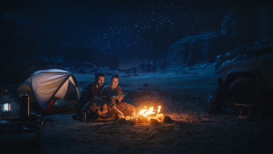 Happy Couple Camping in the Canyon at Night, Use Digital Tablet Computer, Sitting by Campfire. Two people Have Fun, Smile, Post Traveling Photos on Social Media, Watch Funny Videos on Internet