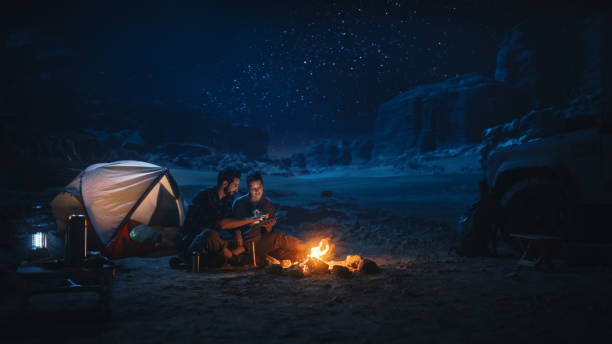istock Happy Couple Camping in the Canyon at Night, Use Digital Tablet Computer, Sitting by Campfire. Two Traveling people Have Fun, Smile, Post on Social Media, Watch Funny Videos on Internet 1330897172