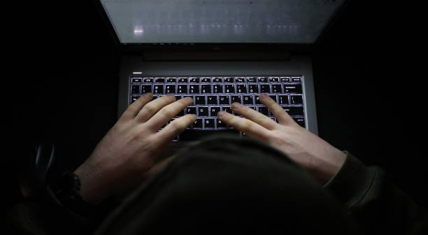 person with a hoody typing at a computer in the dark. suspicious online behavior - computer crime imagens e fotografias de stock