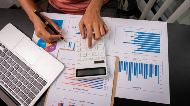 istock Female accountant using calculator and computer holding pen with financial graph and calculate home office expenses on top view table. 1330905887