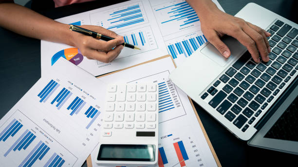 istock Female accountant using calculator and computer holding pen with financial graph and calculate home office expenses on top view table. 1330905900