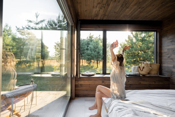 Woman wakes up at country house in nature Woman wakes up in a country house or hotel with panoramic windows in pine forest raised her hands yawning. Good morning and recreation on nature concept hotel window view stock pictures, royalty-free photos & images