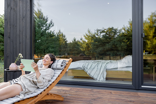 Young woman resting on sunbed and reading on a tablet on terrace at modern house with panoramic windows near pine forest. Concept of solitude and recreation on nature. Wellness and mindful resort