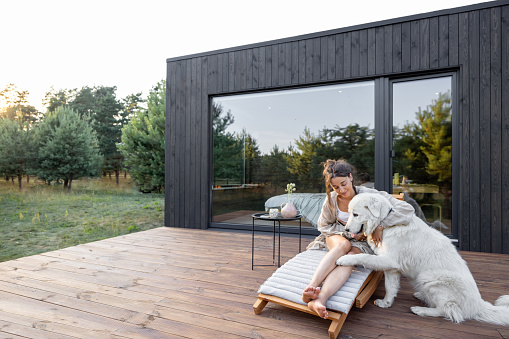 Woman resting on sunbed on wooden terrace near the modern house with panoramic windows near pine forest while playing with pet. Concept of solitude and recreation on nature