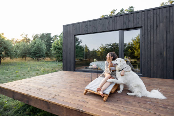 Woman enjoy nature at country house with pet Woman enjoys the nature while sits on sunbed on wooden terrace near the modern house with panoramic windows near pine forest while hugs her pet. Concept of solitude and recreation on nature chalet stock pictures, royalty-free photos & images
