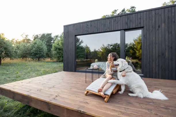 Woman enjoys the nature while sits on sunbed on wooden terrace near the modern house with panoramic windows near pine forest while hugs her pet. Concept of solitude and recreation on nature