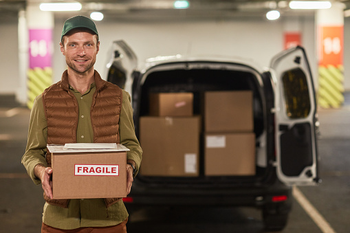 Waist up portrait of male delivery worker holding boxes and smiling at camera while standing by van outdoors, copy space