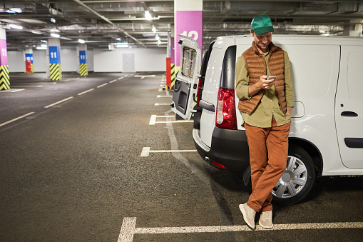 Full length portrait of male delivery worker standing by van at parking lot and using smartphone, copy space