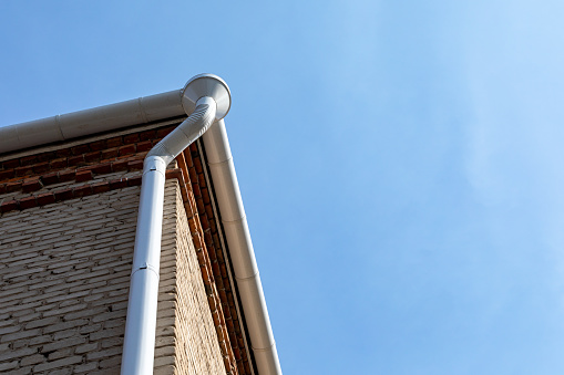 White metal pipe from the drain on the wall of a brick building. Holder gutter drainage system on the roof. Drain on the roof of the house. Roof drainage. Water drainage from the roof.