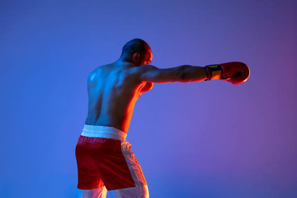 One professional boxer in red shorts training, exercising over purple background in neon light. Back view