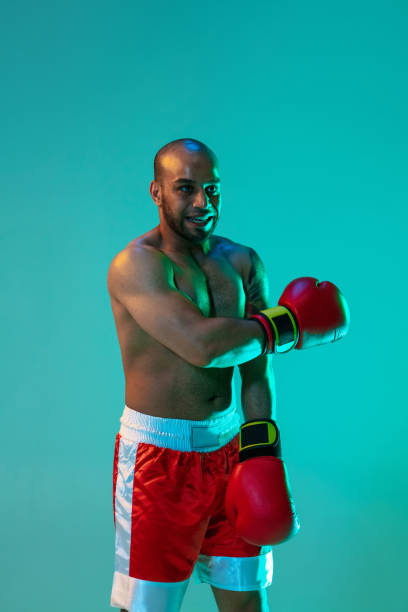 istock Vertical image. One professional boxer in red shorts and gloves posing on blue green background in neon light. 1330922382