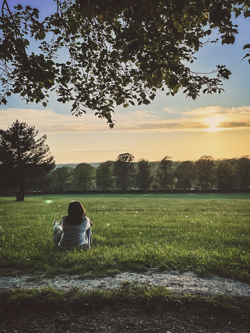 Rear view of a woman with her dog sitting on green field enjoying beautiful sunset