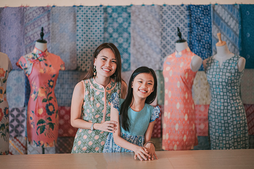 malaysian chinese beautiful mother and daughter portrait in front of her batik fabric textile collection  retail store looking at camera smiling